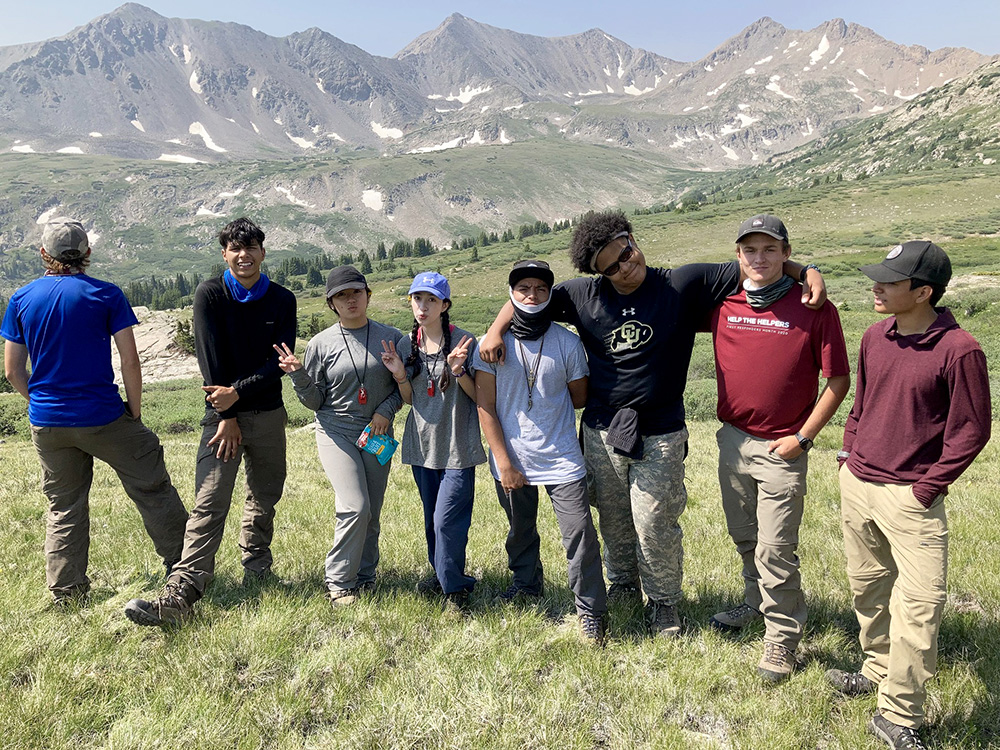 A group stands facing the camera with an alpine valley and mountain peaks in the background.